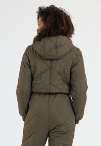 Olivenfärgad vadderad pufferjacka med huva, som har diamantmönster och ribbade mudd, bärs över en matchande jumpsuit.