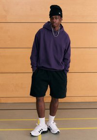 Paarse capuchontrui, zwarte korte broek, witte sportschoenen met zwarte accenten en een zwarte muts. Staand tegen een houten achtergrond.