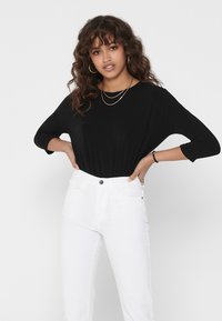 Black long-sleeve shirt with a relaxed fit, paired with high-waisted white jeans. Features a smooth texture and subtle neckline. Gold layered necklaces.