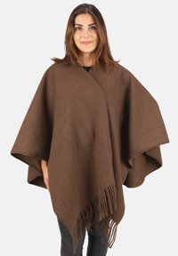 Frau trägt einen braunen Wollponcho mit Fransen, steht vor einem schlichten weißen Hintergrund, blickt frontal und lächelt leicht.