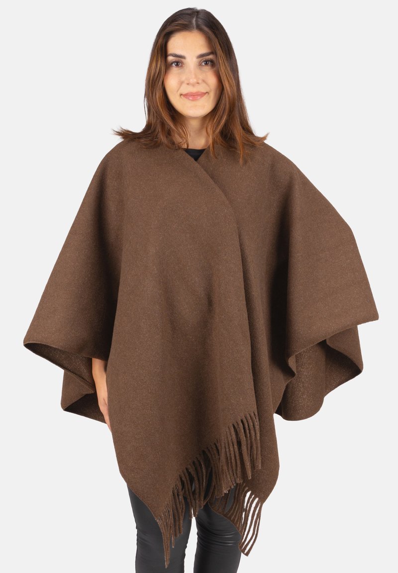 Frau trägt einen braunen Wollponcho mit Fransen, steht vor einem schlichten weißen Hintergrund, blickt frontal und lächelt leicht.