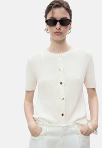 Short-sleeved, white knit cardigan featuring a round neckline and gold button closures down the front; model wears sunglasses and holds pockets.