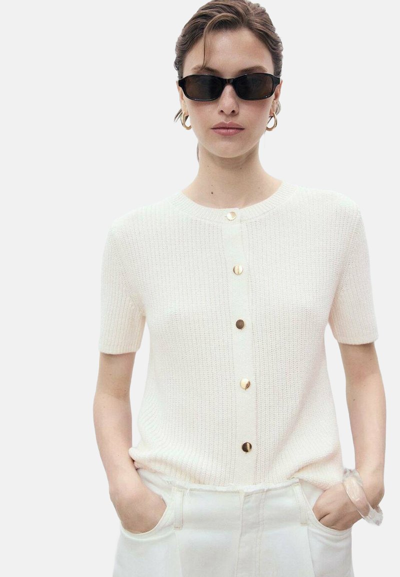 Short-sleeved, white knit cardigan featuring a round neckline and gold button closures down the front; model wears sunglasses and holds pockets.