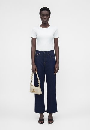 Γυναίκα στέκεται μπροστά σε λευκό φόντο φορώντας λευκό κοντομάνικο πουκάμισο, σκούρο μπλε τζιν, μαύρα σανδάλια, κρατώντας μια μικρή μπεζ τσάντα με δεμένη λουρίδα.