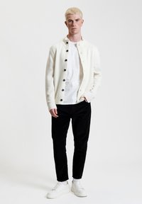 Cardigan bianco lavorato a maglia con bottoni neri, indossato sopra una maglietta bianca, abbinato a pantaloni in corduroy neri e sneakers bianche. Il modello è in piedi contro uno sfondo semplice.