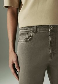 Beige kortemouwen overhemd en lichtgrijze jeans met vijf-pocket ontwerp, gestikte naden en sluiting met een knoop aan de voorkant, vastgehouden door een hand.