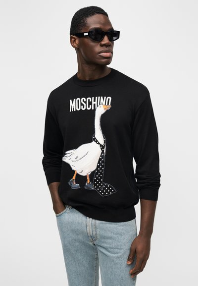 Fekete pulóver liba grafikával, mely pöttyös nyakkendőt visel, felette a "MOSCHINO" logó. A modell sötét napszemüveget és világoskék farmert visel.