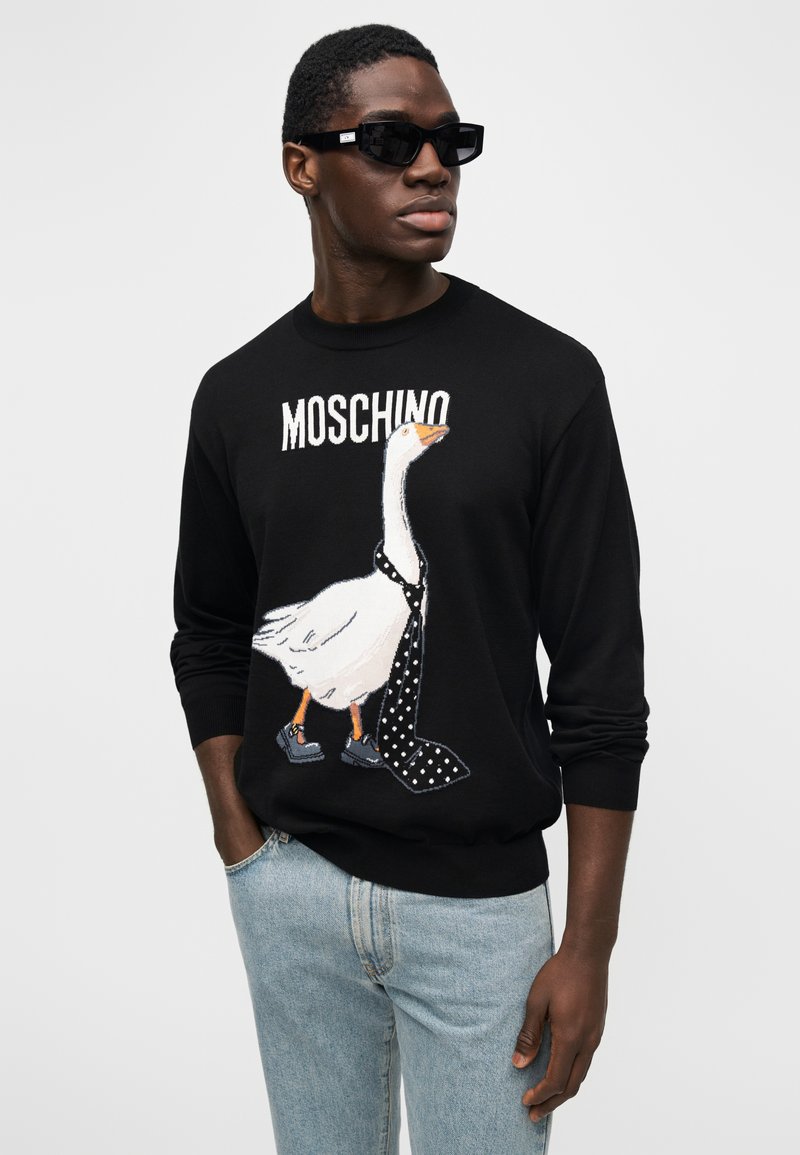 Schwarzer Pullover mit einer Gans-Grafik, die eine gepunktete Krawatte trägt, "MOSCHINO"-Logo darüber. Modell trägt eine dunkle Sonnenbrille und hellblaue Jeans.