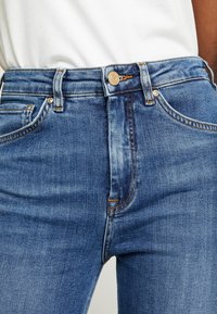 Jeans de mezclilla azul oscuro con cintura alta, que presentan herrajes dorados, bolsillos frontales y detalles de costura amarilla en contraste. Textura suave.