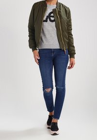 Olivově zelená bomber bunda, šedá mikina s černou grafikou, modré skinny džíny s dírami, černé a růžové tenisky. Jednoduché pozadí.