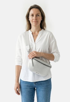 Frau in weißer Bluse und blauen Jeans hält eine silberne Umhängetasche mit strukturierter Oberfläche und Reißverschlussverschluss.