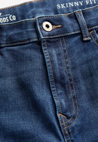 Slim fit denim jeans in donkerblauw met zichtbare stiksels, metalen knoopsluiting en een gestructureerde stof. Merkenlabel bij de tailleband.
