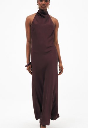 Mujer con un vestido sin mangas, largo hasta el suelo, color púrpura oscuro con cuello halter, grandes pendientes colgantes blancos y una pulsera a juego.