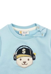 Chemise en coton bleu clair ornée d'un ours brodé portant des écouteurs et une casquette avec la lettre "S". Boutons-pression à l'épaule.