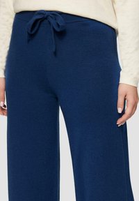 Navyblauwe wijde broek van zachte tricotstof, met een trekkoord in de taille en een gladde textuur. Handen zichtbaar aan de zijkanten.
