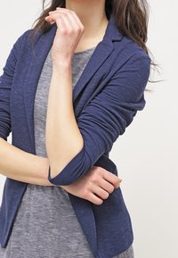 Granatowa marynarka z teksturowanej tkaniny, dopasowany krój i slimowane rękawy, nałożona na szaro-prążkowaną bluzkę. Brak widocznych zapięć.
