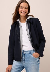 Femme souriante avec de longs cheveux bruns portant une veste bleu marine sur un sweat à capuche gris, un t-shirt blanc et un jean bleu clair, la main dans la poche.