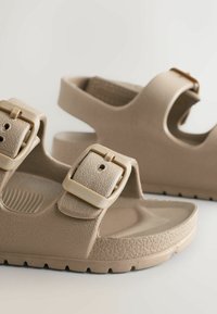 Beige rubberen sandalen met twee verstelbare banden met vierkante gespen, een getextureerd oppervlak, een gevormde voetbed en een patroon op de zool.