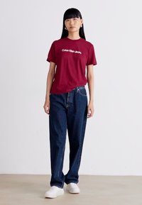 T-shirt bordeaux à manches courtes avec un logo imprimé blanc, accompagné d'un jean en denim foncé ample et de baskets blanches sur un fond uni.
