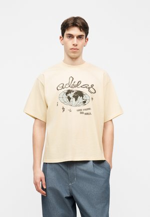 Mladý muž v béžovom veľkom tričku s grafikou glóbusu a textom "Tri pruhy. Jeden svet." v kombinácii s voľnými modrými nohavicami.