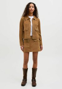 Veste en suède marron avec deux poches avant, assortie à une jupe. Le mannequin porte un haut blanc et des bottes marron mi-mollet. Texture lisse.