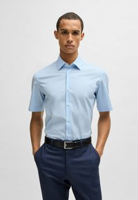 Camicetta azzurra chiara a maniche corte con colletto classico, bottoni frontali e texture liscia, abbinata a pantaloni scuri con motivo e una cintura nera.
