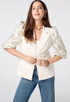 Creme-kleurige blazer met gestructureerd ontwerp en opbollende kantmouwen in lichte groentinten, gecombineerd met blauwe jeans, wat een gelaagde uitstraling creëert.