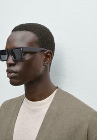 Schwarze rechteckige Sonnenbrille mit dicken Rahmen, getragen von einer Person in einem hellbeigen Hemd und olivgrüner Strickjacke, vor einem blassblauen Hintergrund.