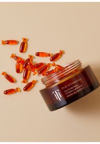 Szklany słoik z bursztynowego szkła z etykietą "True Botanicals Phyto-Retinol Vitamin A Booster" oraz rozsypanymi bursztynowymi kapsułkami żelowymi na beżowym tle.
