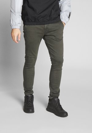 Homme portant un jean skinny olive et des baskets montantes noires, debout avec une main dans la poche devant un fond uni.
