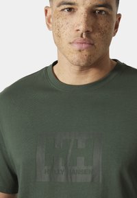 Joven con pecas que lleva una camiseta Helly Hansen verde oliva, mostrado desde el pecho hasta la cara contra un fondo liso.