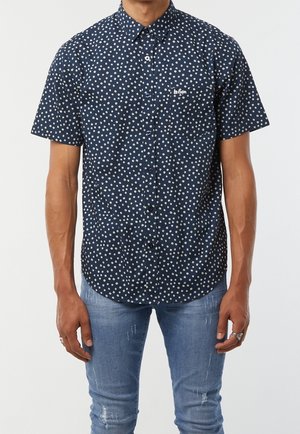 Hombre vistiendo una camisa azul marino de manga corta con botones y lunares blancos, y jeans desgastados de mezclilla azul claro.