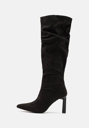 Botte haute noire en suède avec bout pointu et talon carré de hauteur moyenne sur fond gris clair uni.