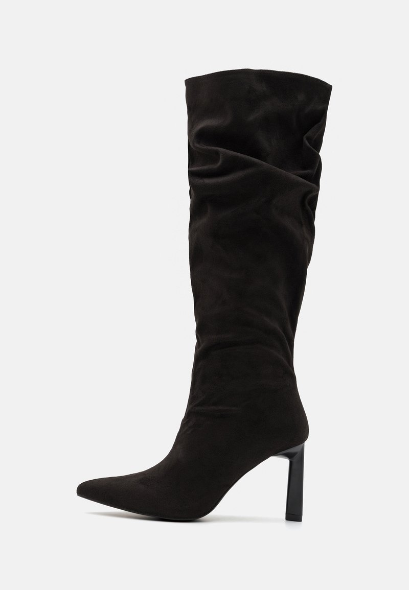 Botte haute noire en suède avec bout pointu et talon carré de hauteur moyenne sur fond gris clair uni.