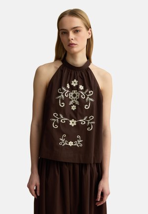 Femme portant un haut sans manches marron foncé avec broderie florale blanche, associé à une jupe marron foncé assortie.