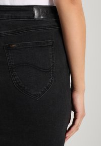 Falda de mezclilla negra con un diseño de bolsillo trasero curvado y una etiqueta de cuero en la cinturilla. Textura suave y forma ajustada.