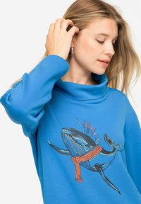 Sudadera azul con cuello de cowl, que presenta un gráfico de una ballena que lleva una bufanda naranja. Hecha de una tela suave y lisa con mangas largas.
