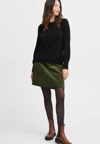 Schwarzer Strickpullover mit langen Ärmeln, kombiniert mit einem dunkelgrünen Cordrock. Das Model trägt schwarze Strumpfhosen und braune Stiefeletten.