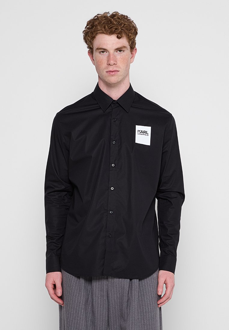 Chemise noire à manches longues en tissu lisse et brillant avec un col boutonné. Présente un patch logo blanc sur la poitrine gauche et des boutons en métal.