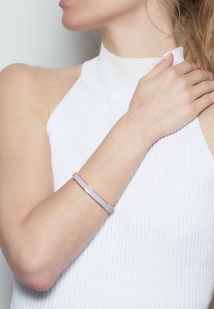Thin, rose gold bangle bracelet with a smooth, glossy white accent. Worn on a bare wrist, paired with a high-neck white ribbed top.