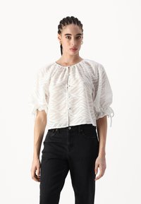 Bluză albă, semi-transparentă, cu un model ondulat, guler strâns, și mâneci scurte tip puff, asociată cu blugi negri din denim. Botoane pe toată lungimea din față.