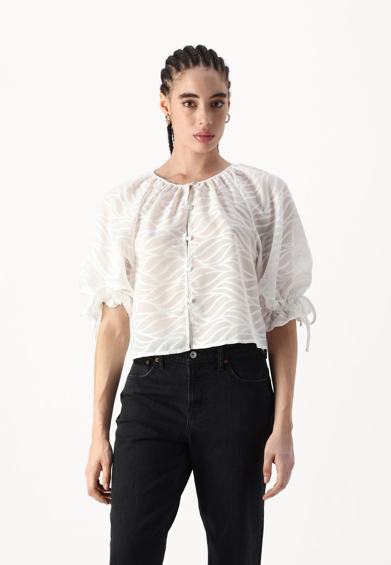 Bluză albă, semi-transparentă, cu un model ondulat, guler strâns, și mâneci scurte tip puff, asociată cu blugi negri din denim. Botoane pe toată lungimea din față.