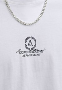 Biele tričko s textom „Mission Statement CORE CREATIVE DEPARTMENT“ a logom vavrínového venca, doplnené o strieborný retiazkový náhrdelník.