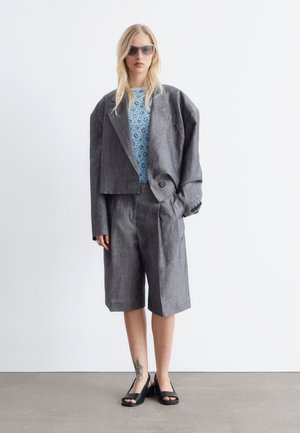 Lyshåret kvinde iført oversized grå blazer, matchende lange shorts, blå blomster top, sorte åbne hæle, solbriller, stående på gråt gulv.