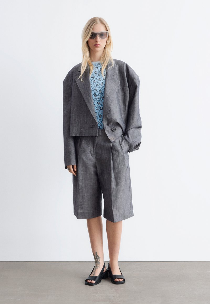 Femme blonde portant un blazer gris surdimensionné, un short long assorti, un haut bleu à fleurs, des talons ouverts noirs, des lunettes de soleil, debout sur un sol gris.