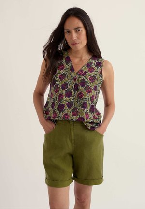 Mujer con el cabello largo y oscuro, vestida con una blusa floral sin mangas y pantalones cortos verde oliva con dobladillo enrollado, de pie con las manos en los bolsillos frente a un fondo liso.