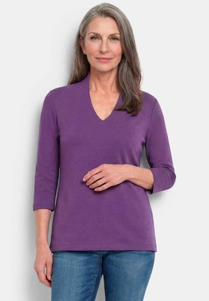 Femme aux cheveux gris portant un haut violet à col en V avec des manches trois-quarts et un jean bleu, debout devant un fond clair uni.