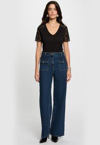 Schwarzes V-Ausschnitt-Oberteil mit Spitzenärmel, kombiniert mit dunkelblauen High-Waist-Breitbeinjeans mit Fronttaschen und gesteppten Details.