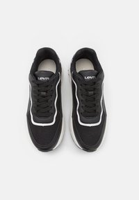 Sorte sneakers med en teksturert overdel, hvite detaljer, snøring og en kraftig såle. Merkenavnet er synlig på tungen.