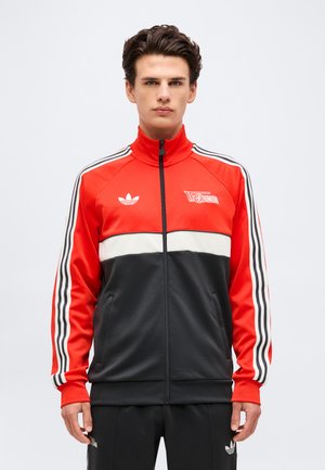 Młody mężczyzna w czerwono-czarno-białej kurtce dresowej Adidas z logo 1. FC Union Berlin, stojący na jednolitym tle.